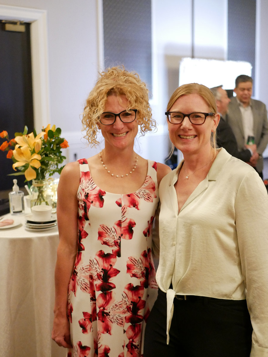 Two blonde women wearing glasses (one with tight curls, the other with straight hair) stand next to each other. Behind them is a tall table with a white table cloth. On the table is a bouquet of orange flowers like lillies. 